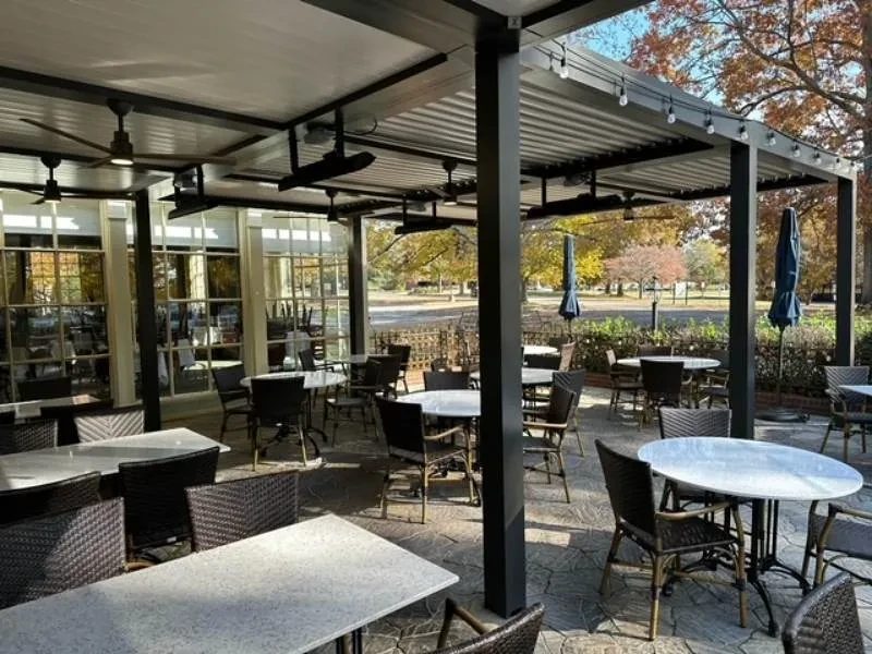 Outdoor restaurant patio with dark pergola, seating, and autumn trees.