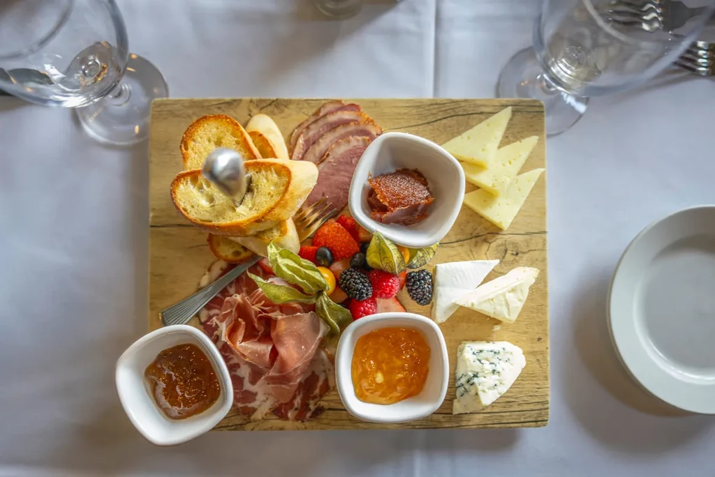 Top-down charcuterie and cheese board with meats, cheeses, fruits, and baguette.