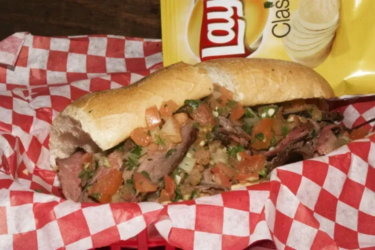 A steak sandwich with salsa in a basket, with a chip bag.