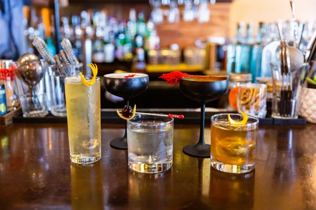 Four garnished cocktails and one clear drink on a wooden bar.