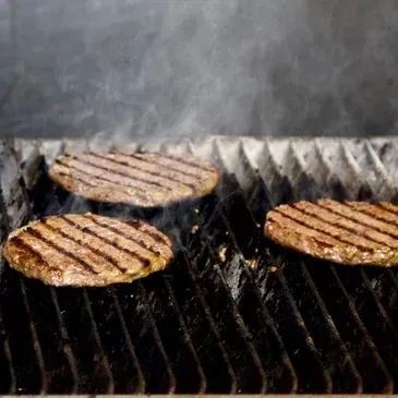 Three hamburger patties cook on a black grill, producing smoke.