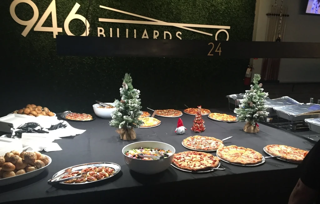 A black table laden with pizzas, salads, and bread, adorned with mini Christmas trees.