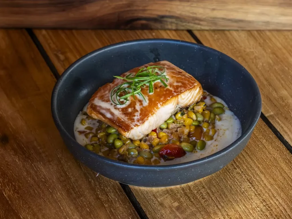 Seared salmon with succotash and sauce in a black bowl on wood.