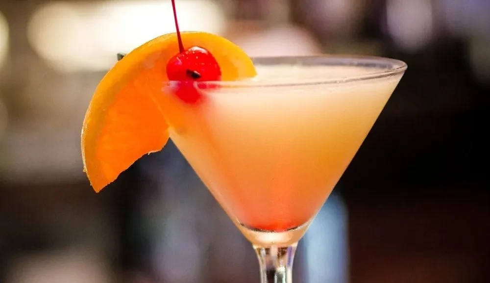 A peach-colored cocktail with an orange slice and cherry garnish.