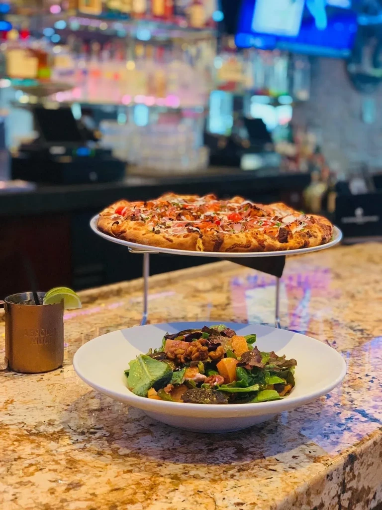 A pizza on a stand above a salad and drink on a bar.