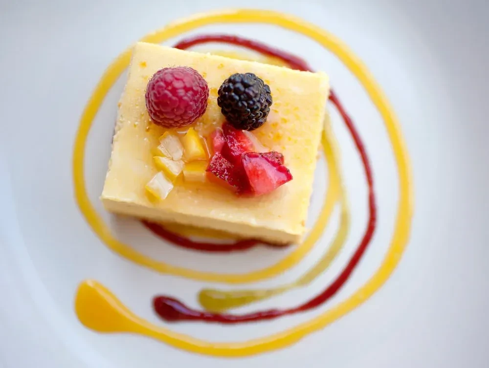 A slice of cheesecake with berries and fruit, on a decorated white plate.