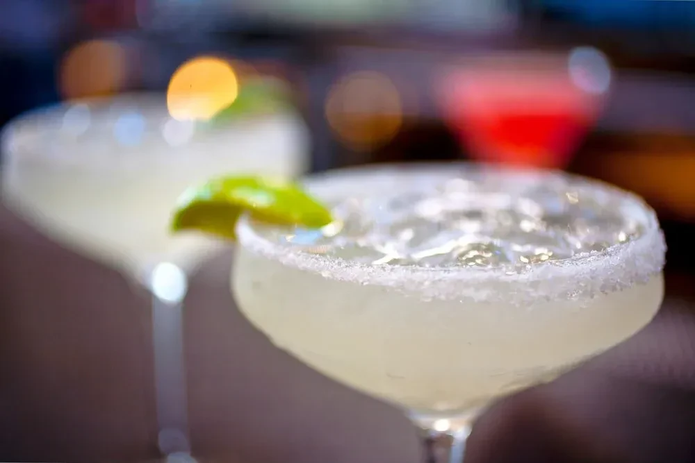 Two margaritas with salt rims and lime wedges, one in focus.
