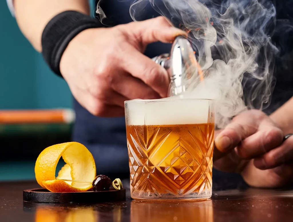 Bartender smoking an Old Fashioned cocktail with an orange peel garnish.