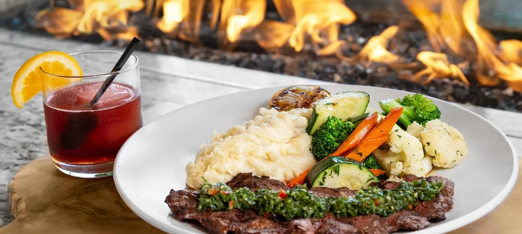 Steak, mashed potatoes, and vegetables with a drink next to a fire.