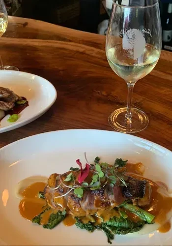Two plates of gourmet food and two glasses of white wine on a wooden table.