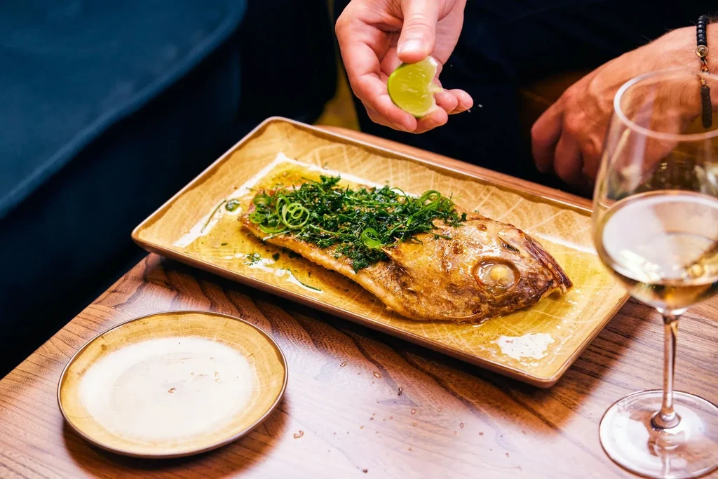 Hand squeezing lime over whole roasted fish on a plate with wine.