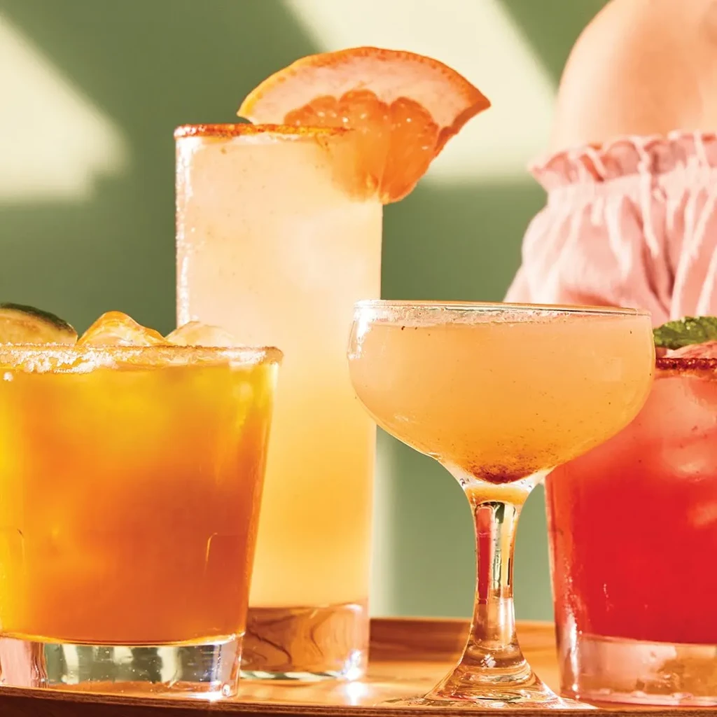 Four colorful alcoholic drinks, garnished, sit on a wooden tray.