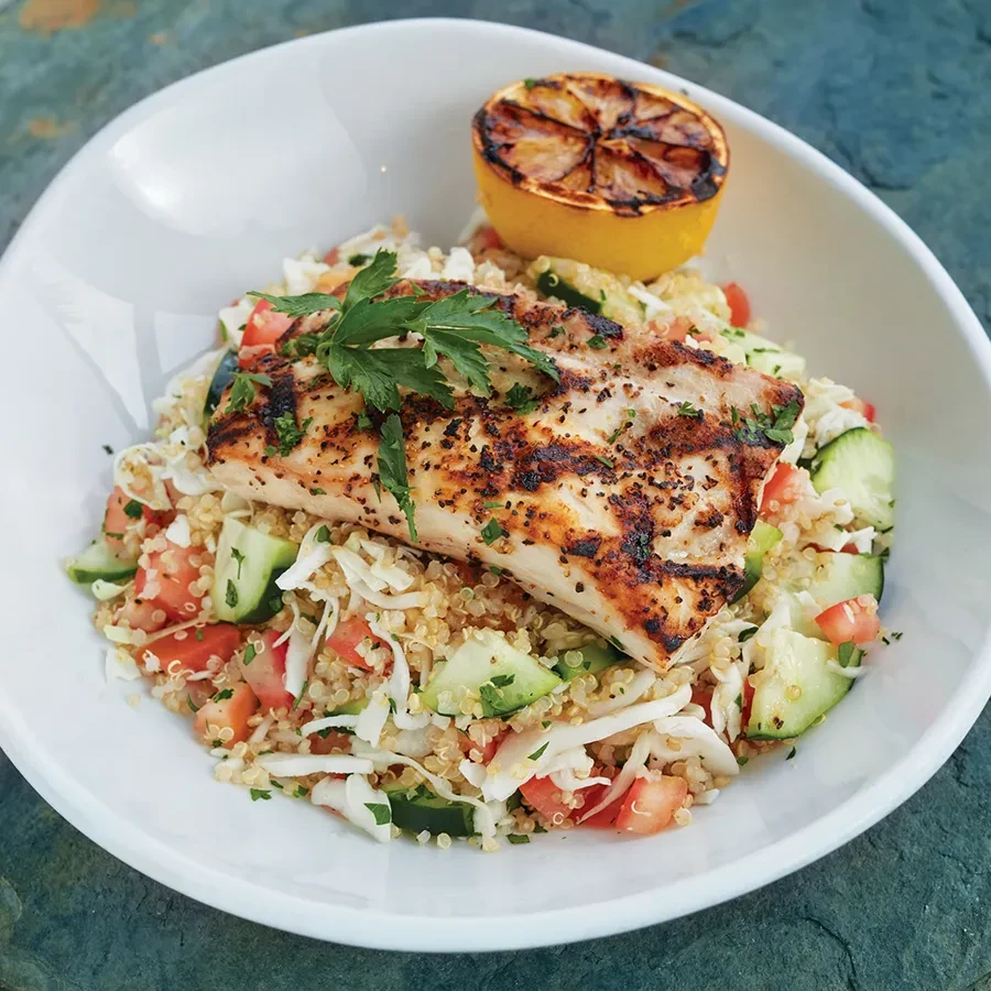 Grilled fish with quinoa salad, cucumber, tomato, and a charred lemon.