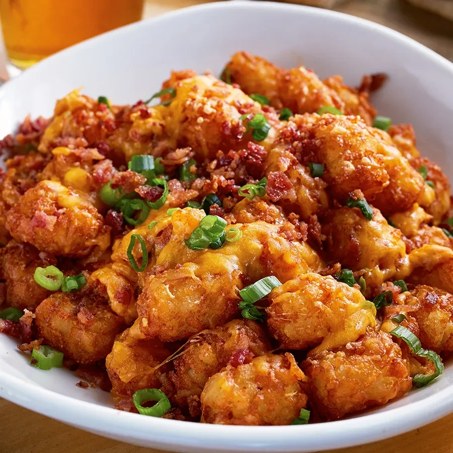 A bowl of golden tater tots topped with cheese, bacon bits, and chives.