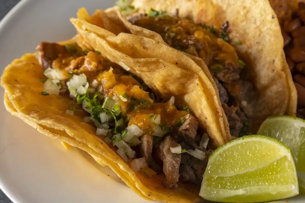 Three street tacos with meat, onions, cilantro, salsa, and lime wedges.