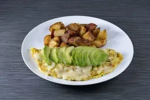 A plate with an omelet topped with avocado slices and a side of roasted potatoes.