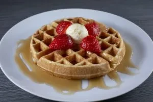 A golden waffle topped with strawberries, cream, and syrup on a white plate.