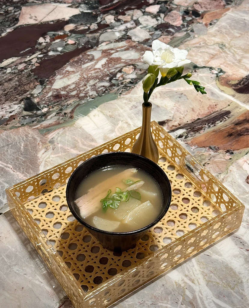 Soup with meat and vegetables, a flower in a vase on a rattan tray.