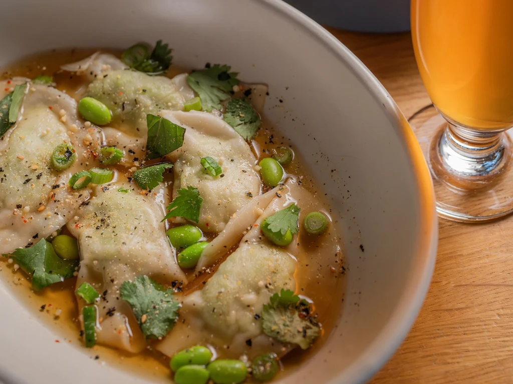 A white bowl of dumplings in broth with edamame and herbs.