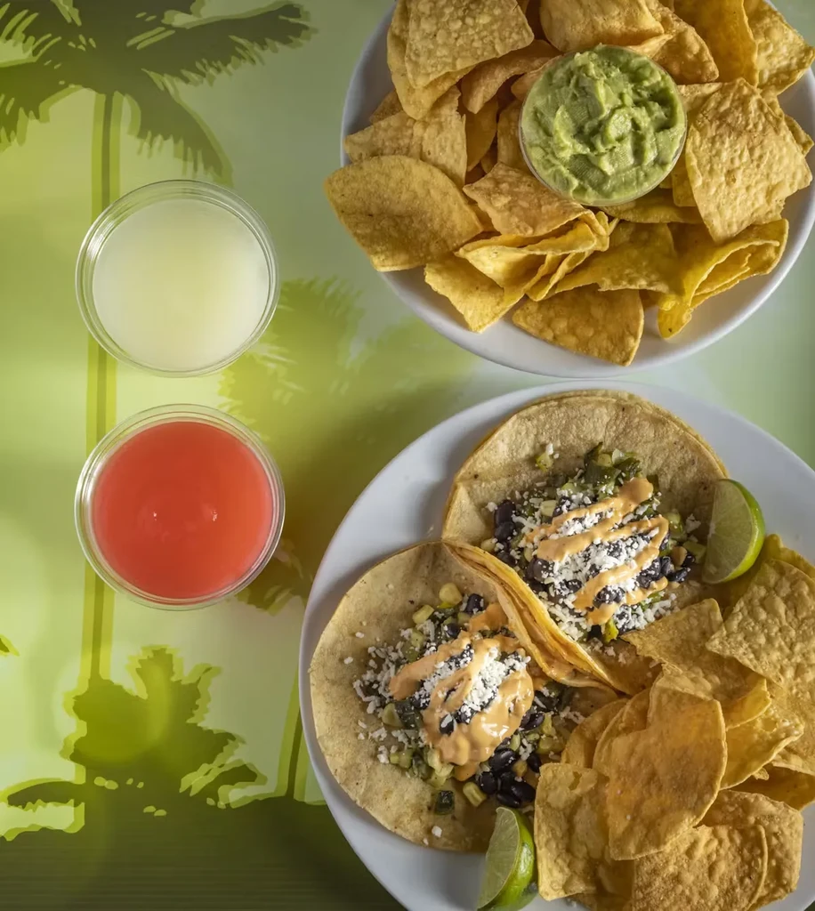 Tacos, chips with guacamole, and two drinks on a palm-tree mat.