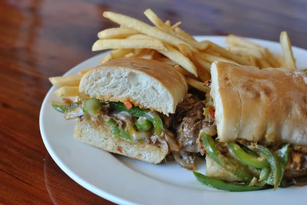 A steak sandwich with green peppers, onions, and fries on a white plate.