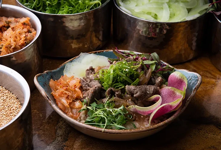 A colorful bulgogi beef rice bowl with various Korean side dishes.