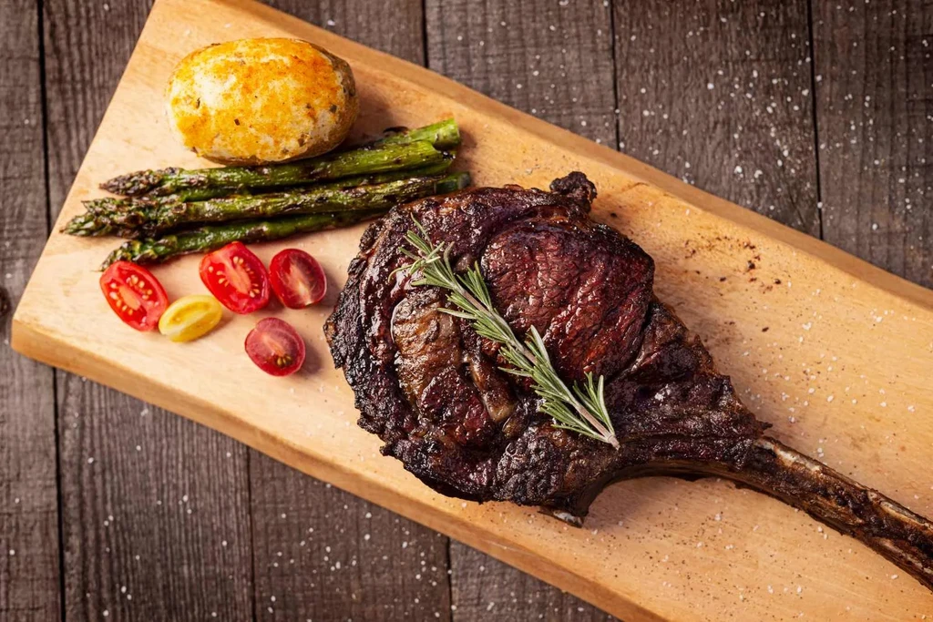 Tomahawk steak with asparagus, cherry tomatoes, and a bun on a wooden board.