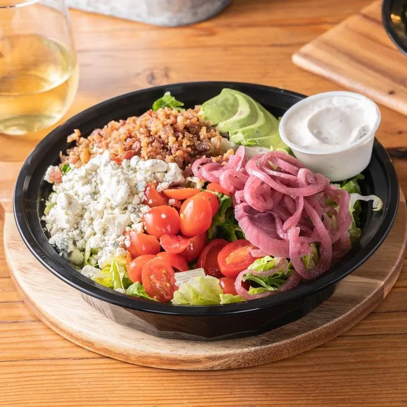 Black bowl of salad with bacon, blue cheese, tomatoes, avocado, and pickled onions.
