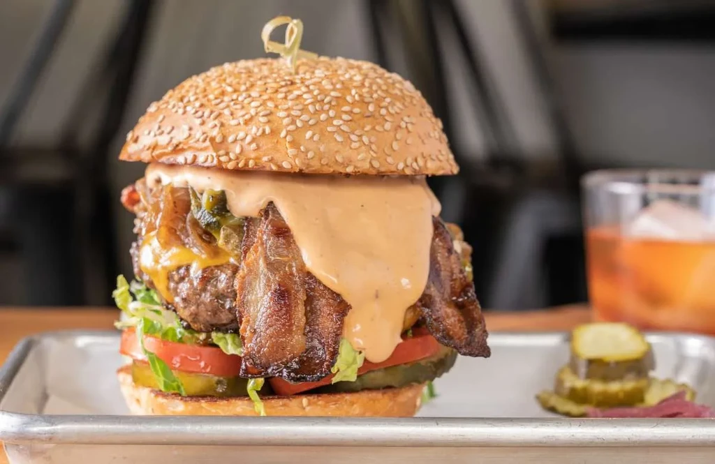 A tall bacon cheeseburger with sauce and a side of pickles on a tray.