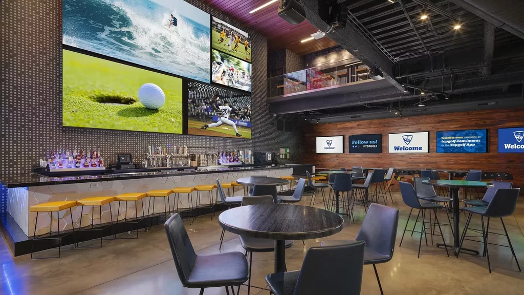 Spacious Topgolf bar with large sports screens and yellow hexagonal stools.