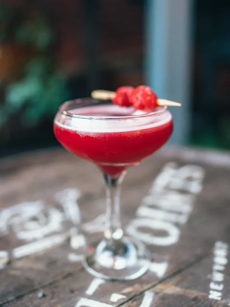 A red cocktail in a coupe glass, garnished with raspberries.