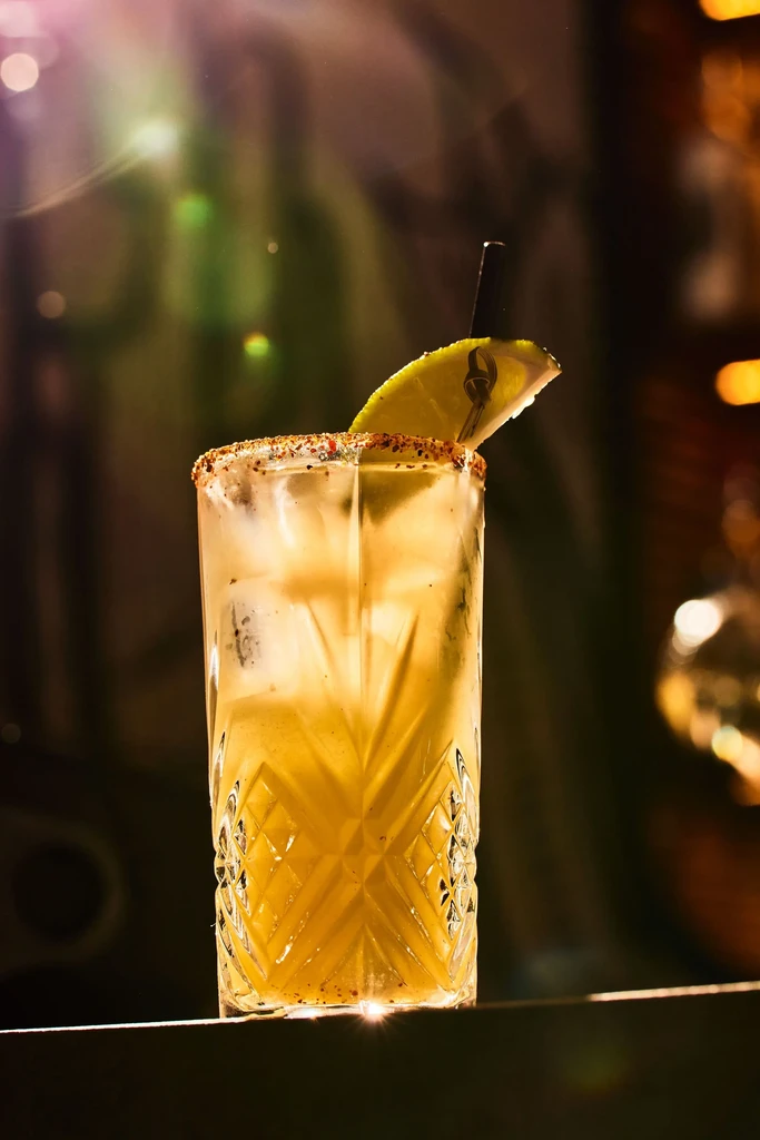 A yellow cocktail with a lime wedge and spiced rim in a patterned glass.
