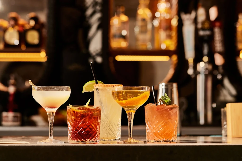 Five cocktails on a bar counter with blurred liquor bottles in background.