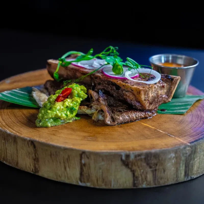 Grilled short ribs with guacamole, onions, and greens on a wooden board.