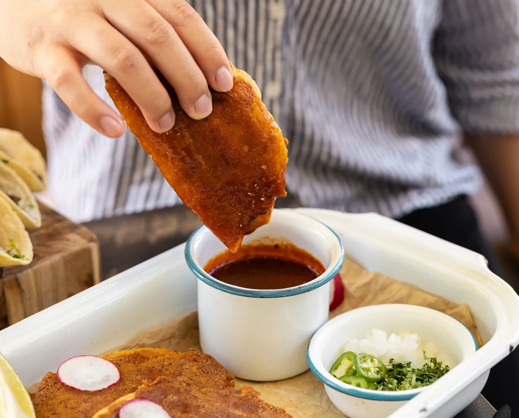 A hand dips a crispy taco into a small cup of dark red sauce.