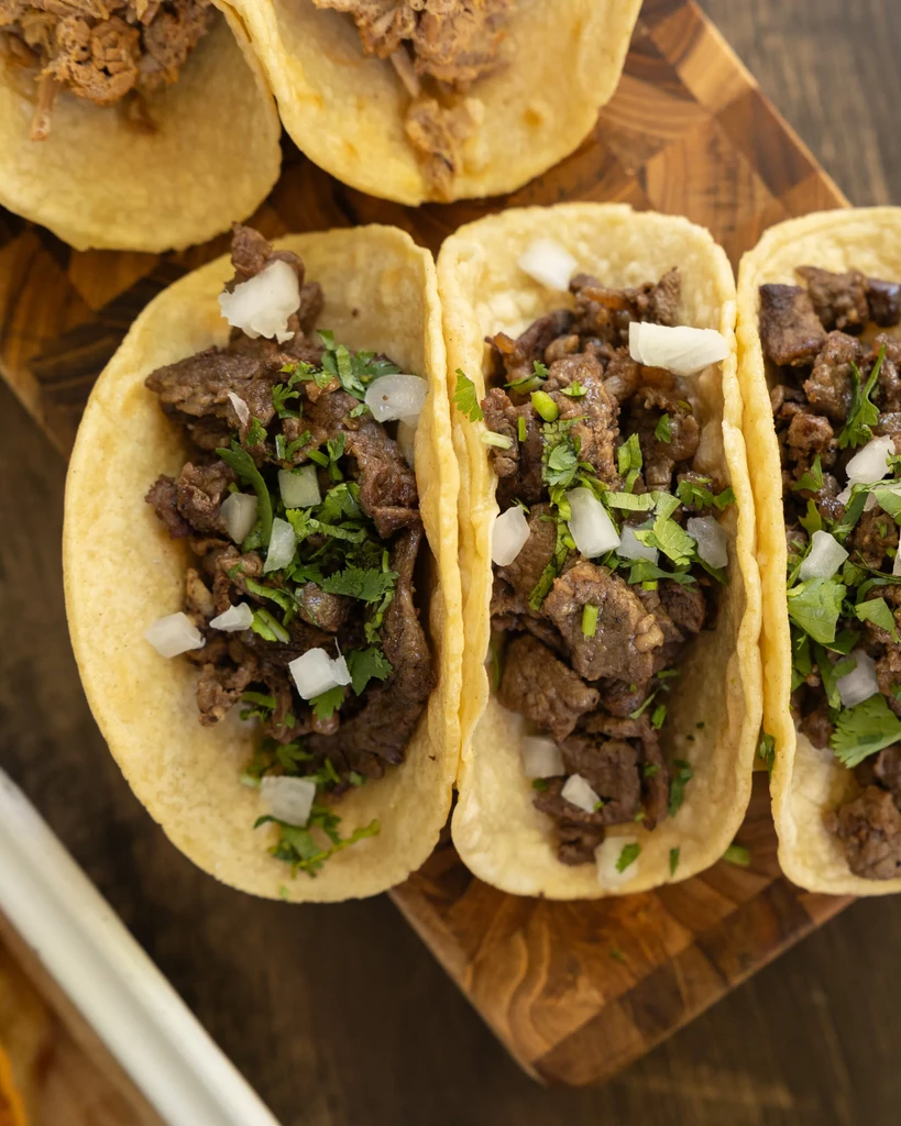 Three beef tacos with onion and cilantro on a wooden board.