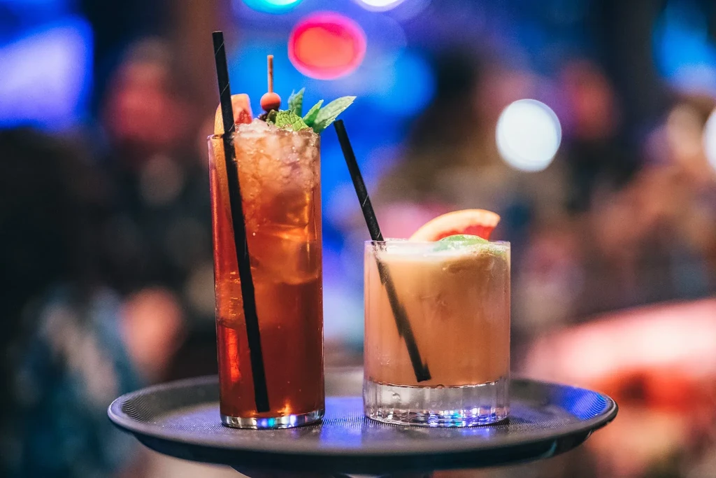 Two garnished cocktails on a black tray against a blurred, colorful background.
