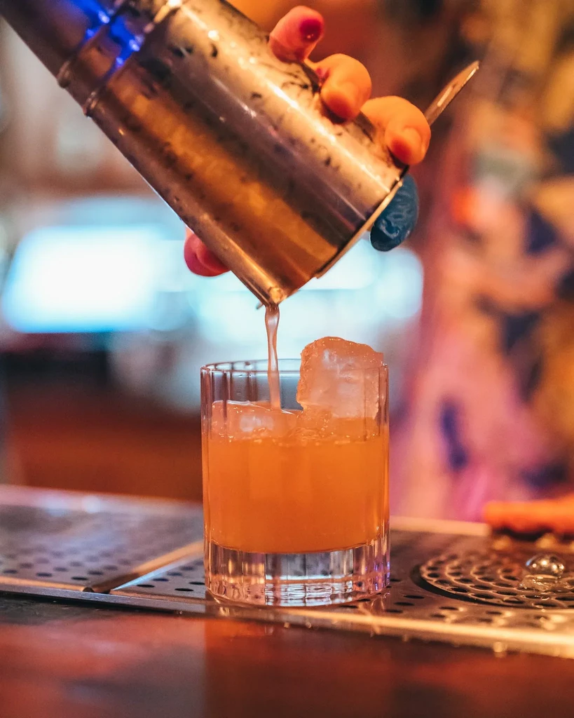 A person pouring an orange cocktail from a shaker into a glass with ice.