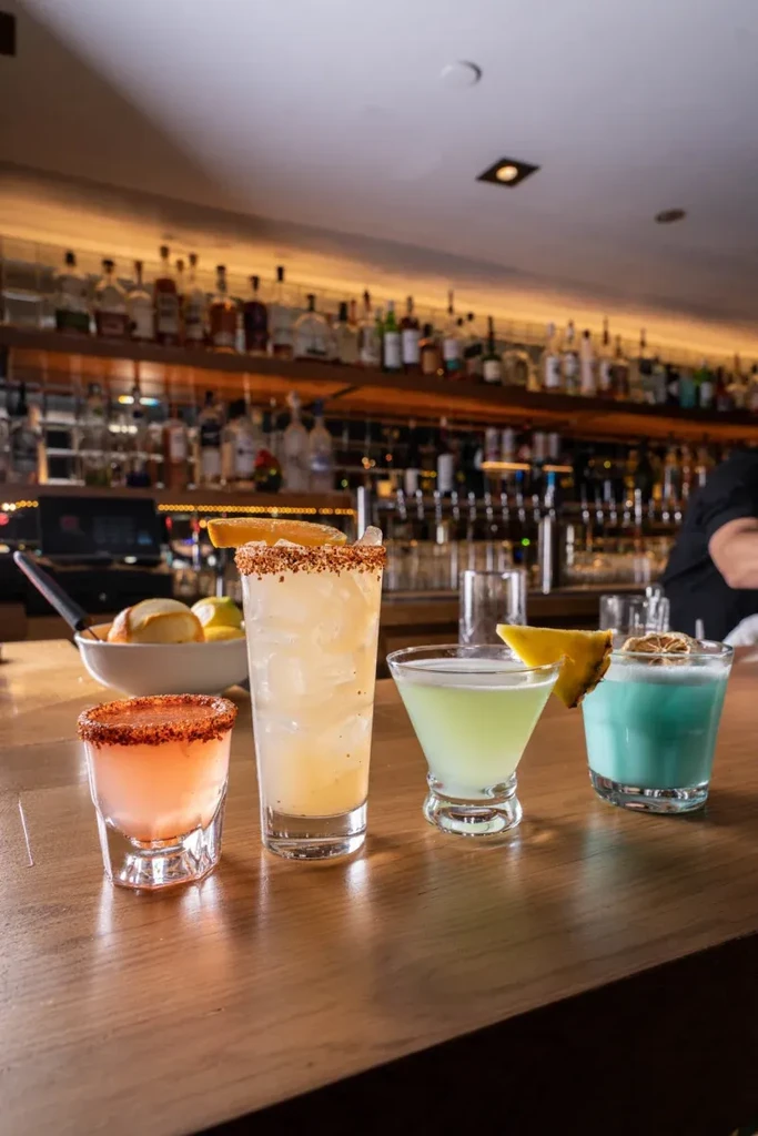 Four colorful cocktails and a shot glass on a wooden bar counter.
