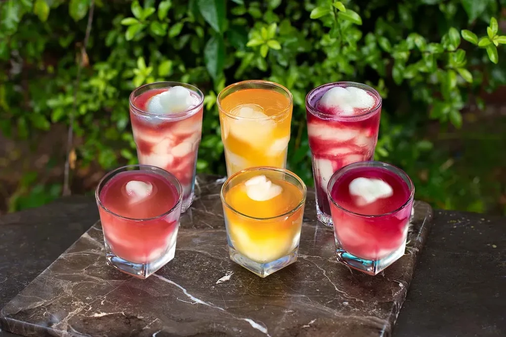 Six colorful drinks with white foam on a marble slab, green background.