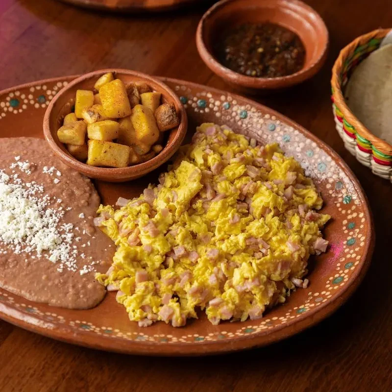 Mexican breakfast platter with scrambled eggs, ham, beans, potatoes, and tortillas.