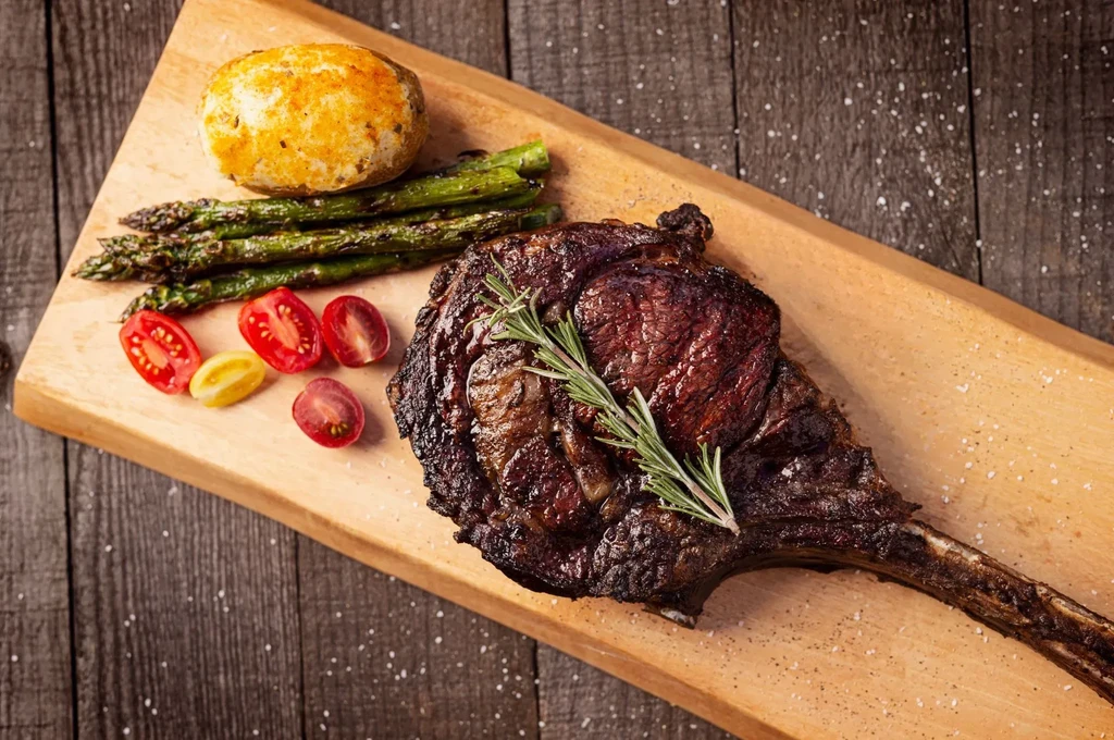 Tomahawk steak with rosemary, asparagus, potatoes, and tomatoes on a wooden board.