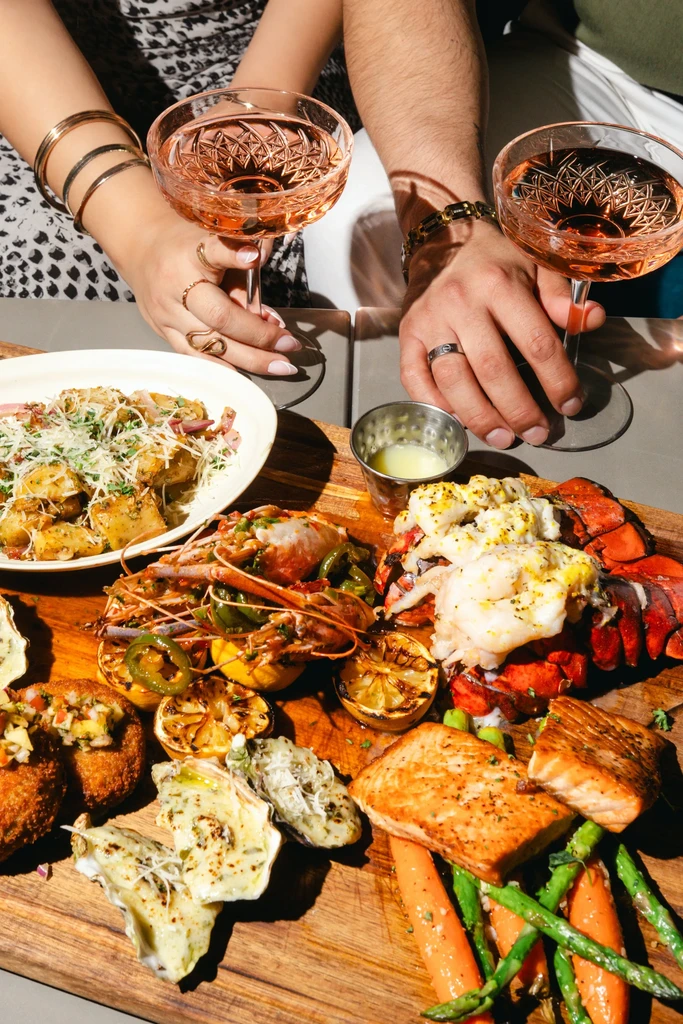 Two people holding cocktails with seafood, pasta, and vegetables on a wooden board.
