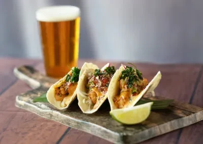Three tacos with a lime wedge on a cutting board, with a beer.