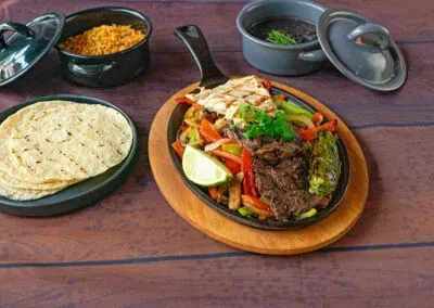Skillet of fajitas with tortillas, rice, and beans on a wooden table.