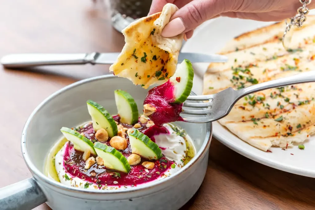 A hand holds flatbread while a fork serves beet dip and cucumber.