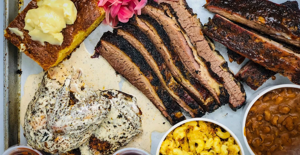 A barbecue platter with brisket, ribs, corn cake, chicken wings, mac-n-cheese, and baked beans.