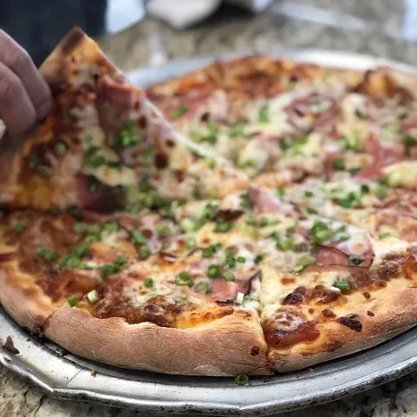 Hand taking a slice of pizza topped with ham and green onions.