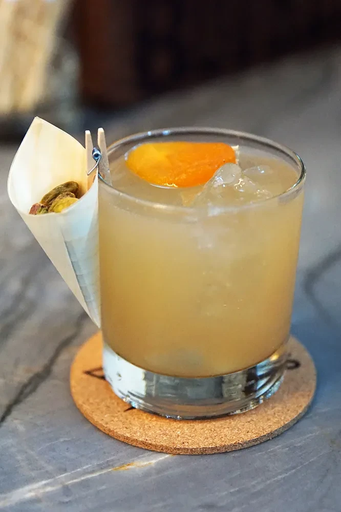 Cocktail with orange slice and pistachios in a cone on a cork coaster.