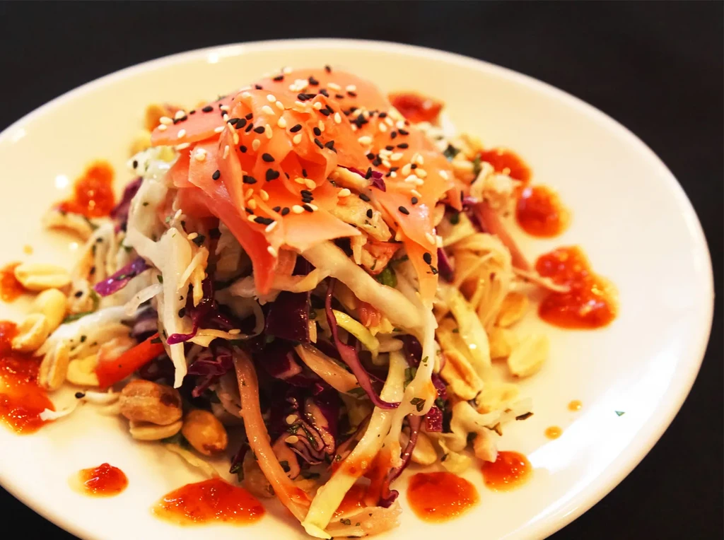 A white plate with a colorful salad, peanuts, and red sauce.